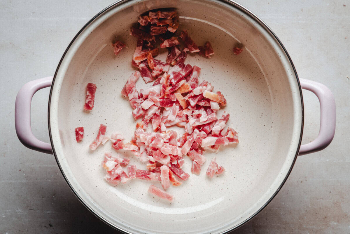 Chopped raw bacon pieces are spread in a large, empty beige pot with purple handles, set on a light-colored surface.