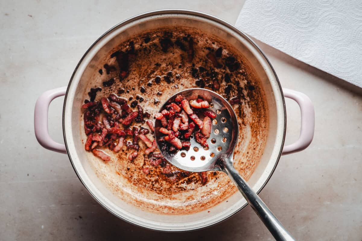 A large pot with browned bits and cooked bacon pieces being scooped up with a slotted spoon. A sheet of paper towel sits nearby on the countertop.