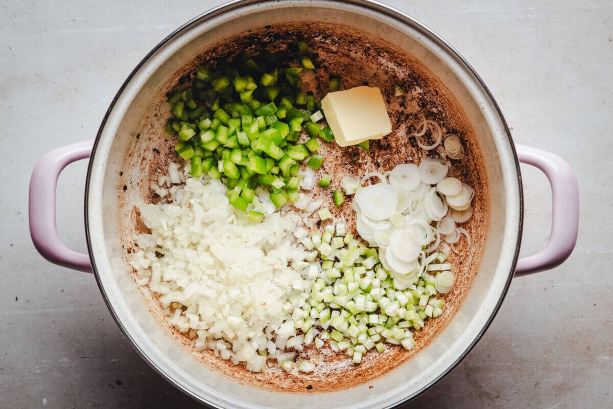 A large pot with chopped celery, green bell pepper, onions, and sliced green onion inside, along with a pat of butter, ready to be cooked. The pot has light purple handles and sits on a light-colored surface.