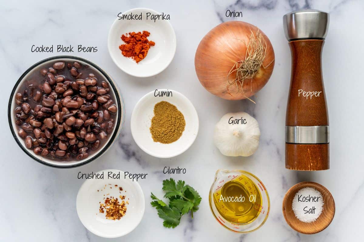 Ingredients for a recipe laid out on a white surface: cooked black beans, smoked paprika, onion, cumin, pepper, garlic, kosher salt, avocado oil, cilantro, and crushed red pepper. Each item is labeled.