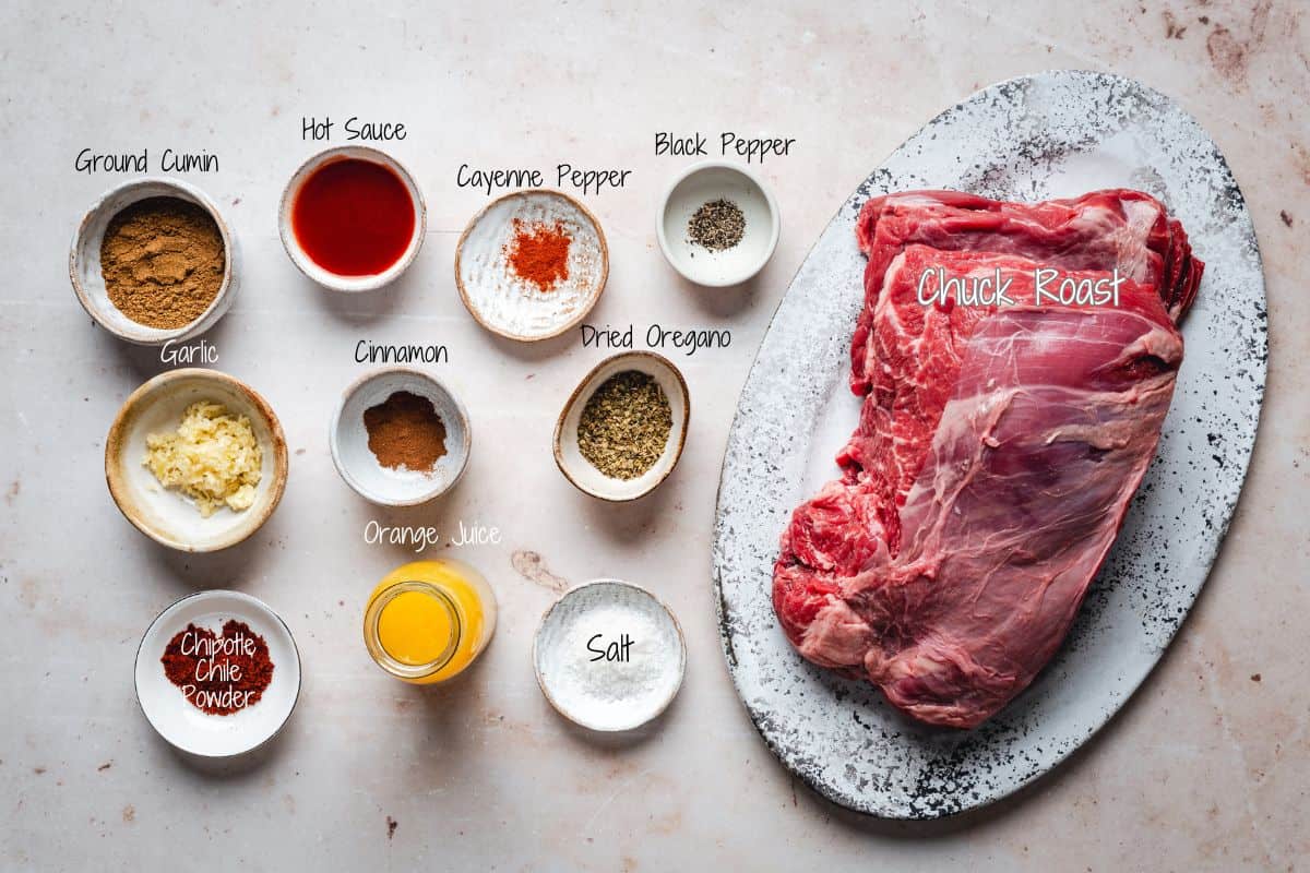 A raw chuck roast on a white oval plate is surrounded by small bowls containing ground cumin, hot sauce, cayenne pepper, black pepper, garlic, cinnamon, dried oregano, chipotle chile powder, orange juice, and salt.