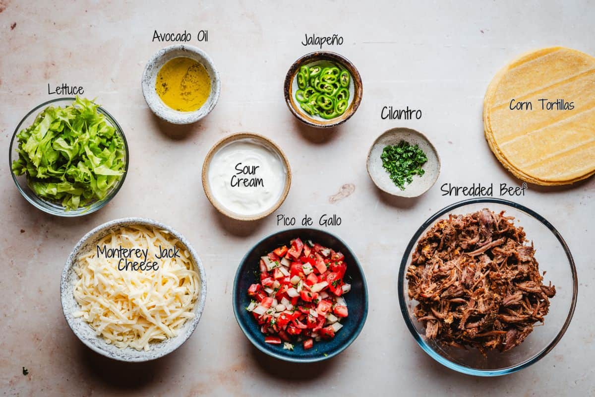 Overhead view of taco ingredients in bowls: shredded lettuce, Monterey Jack cheese, avocado oil, sour cream, pico de gallo, sliced jalapeño, chopped cilantro, shredded beef, and a stack of corn tortillas.