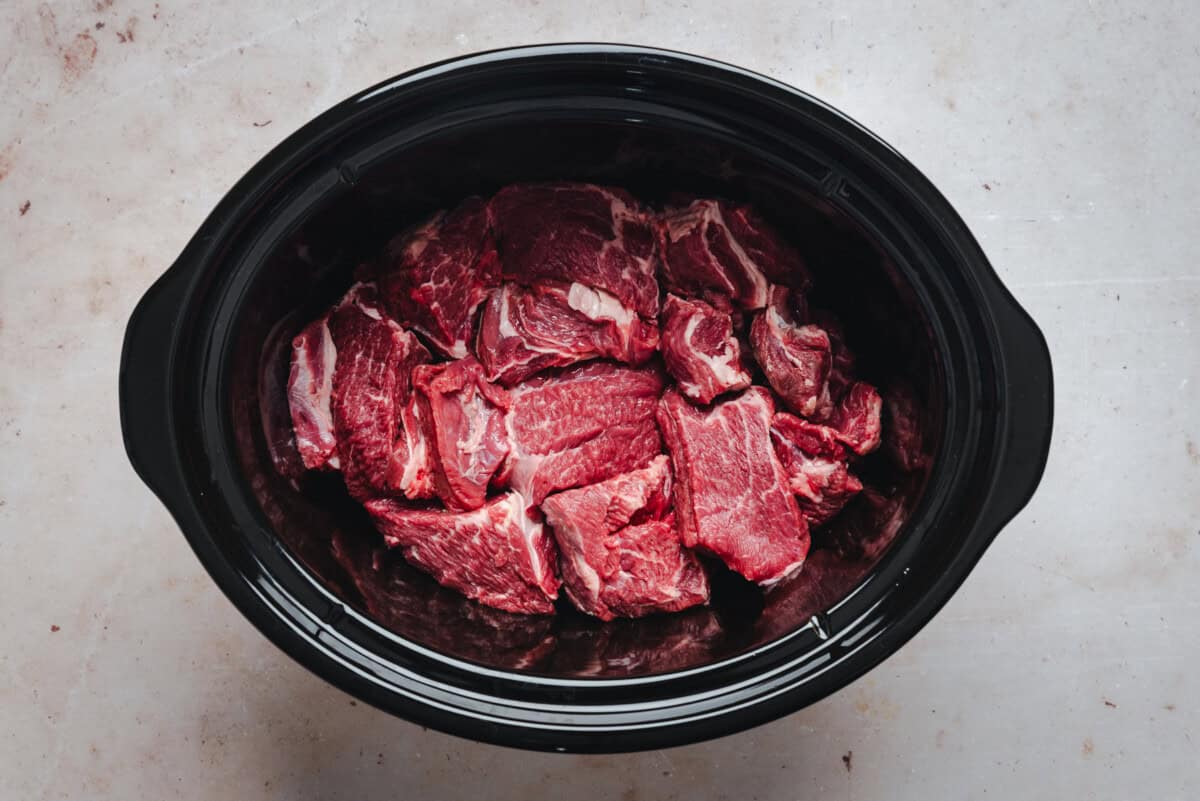 A black slow cooker filled with raw, cubed pieces of red beef, placed on a light-colored countertop.
