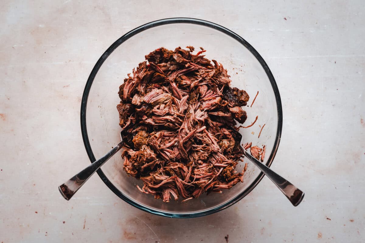 A glass bowl filled with shredded beef, with two metal forks resting inside, placed on a light-colored surface.