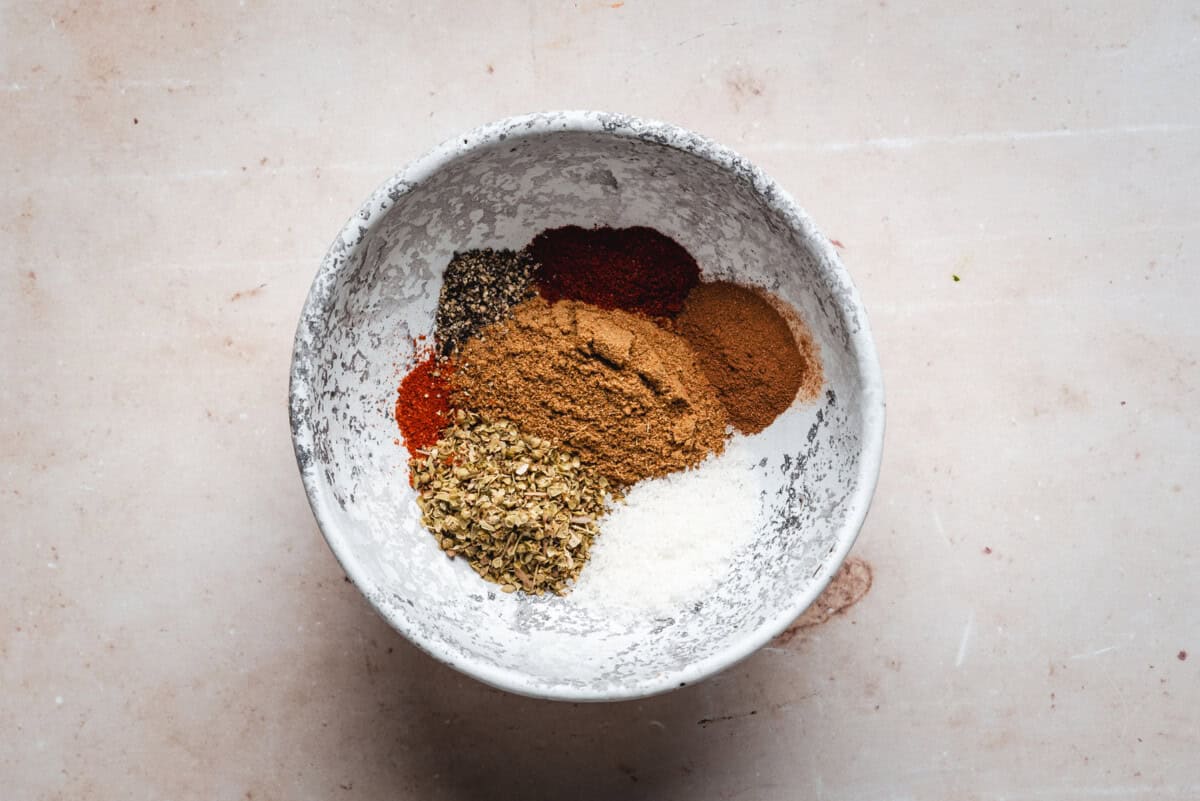 A white bowl on a light surface filled with assorted ground spices, including black pepper, smoked paprika, cinnamon, dried oregano, and salt, arranged in separate piles.