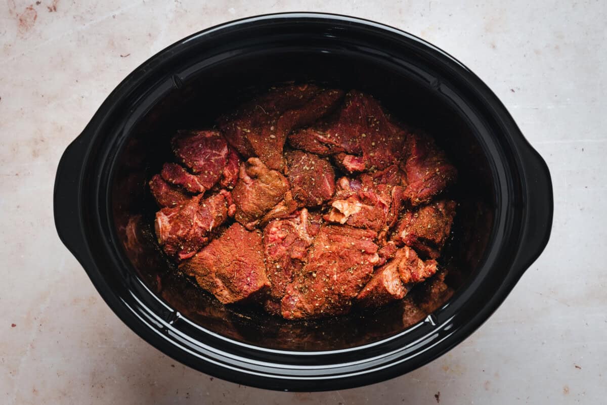 Chunks of raw, seasoned beef sit inside a black slow cooker on a light-colored surface, ready to be cooked.