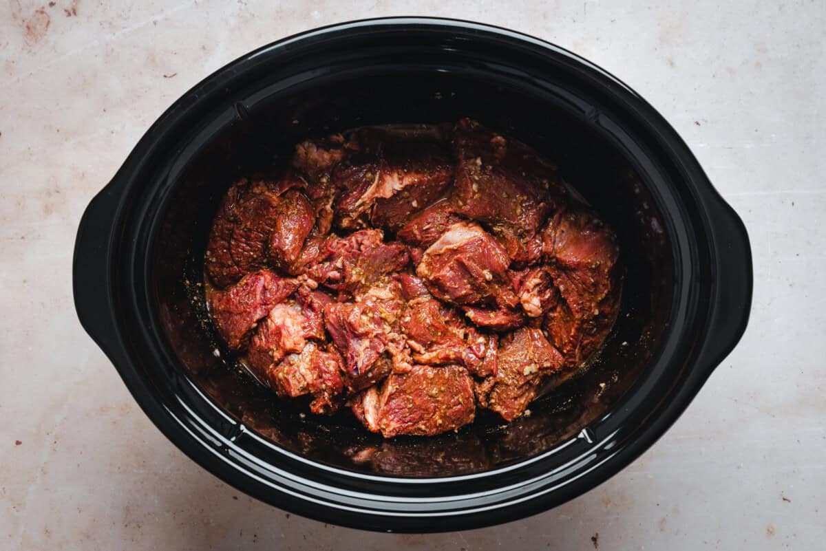 Chunks of raw, seasoned beef are placed inside a black slow cooker on a light-colored countertop, ready to be cooked.