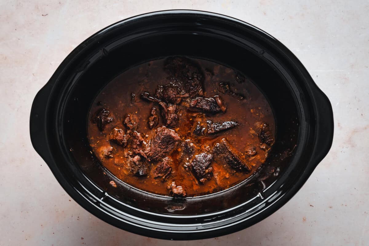 A black slow cooker filled with chunks of cooked beef in a rich, brown sauce, sitting on a light-colored surface.