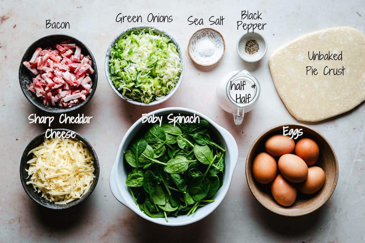 A top-down view of ingredients for spinach bacon quiche, including bowls of chopped bacon, shredded cheddar cheese, sliced green onions, spinach, sea salt, black pepper, half & half, whole eggs, and an unbaked pie crust.