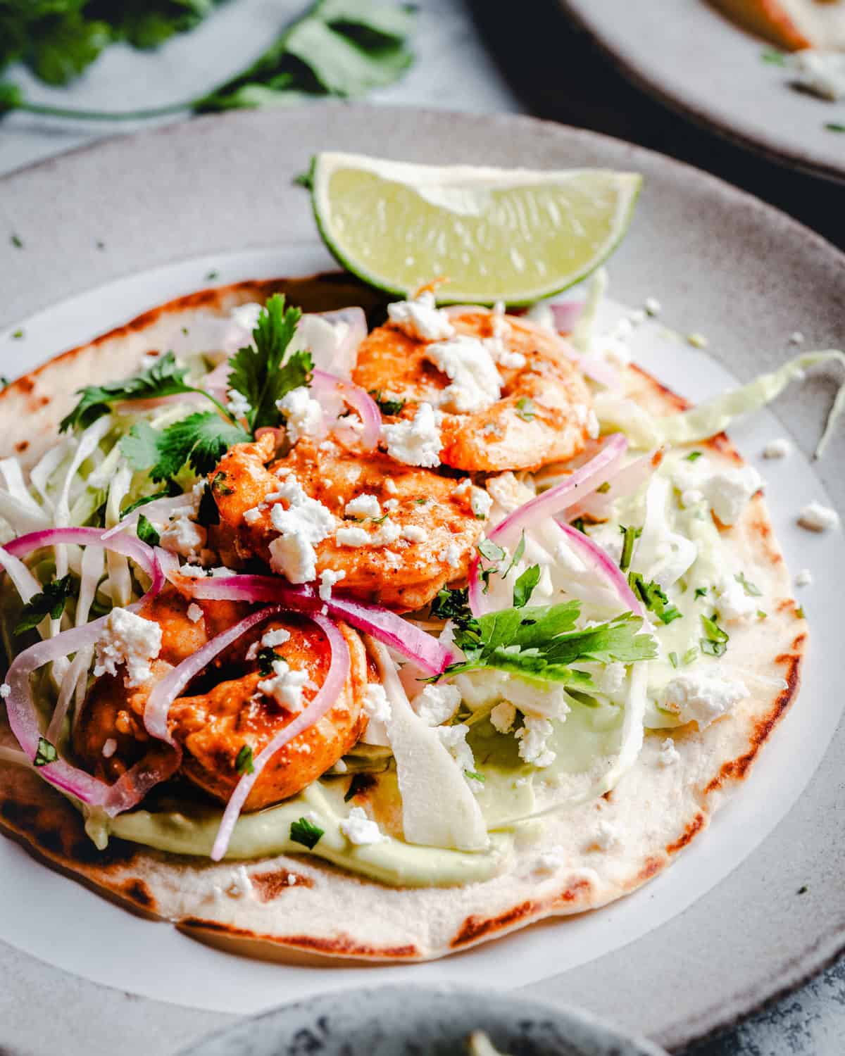 A tostada topped with shrimp, shredded lettuce, pickled red onions, crumbled cheese, fresh cilantro, and a lime wedge, served on a gray plate.