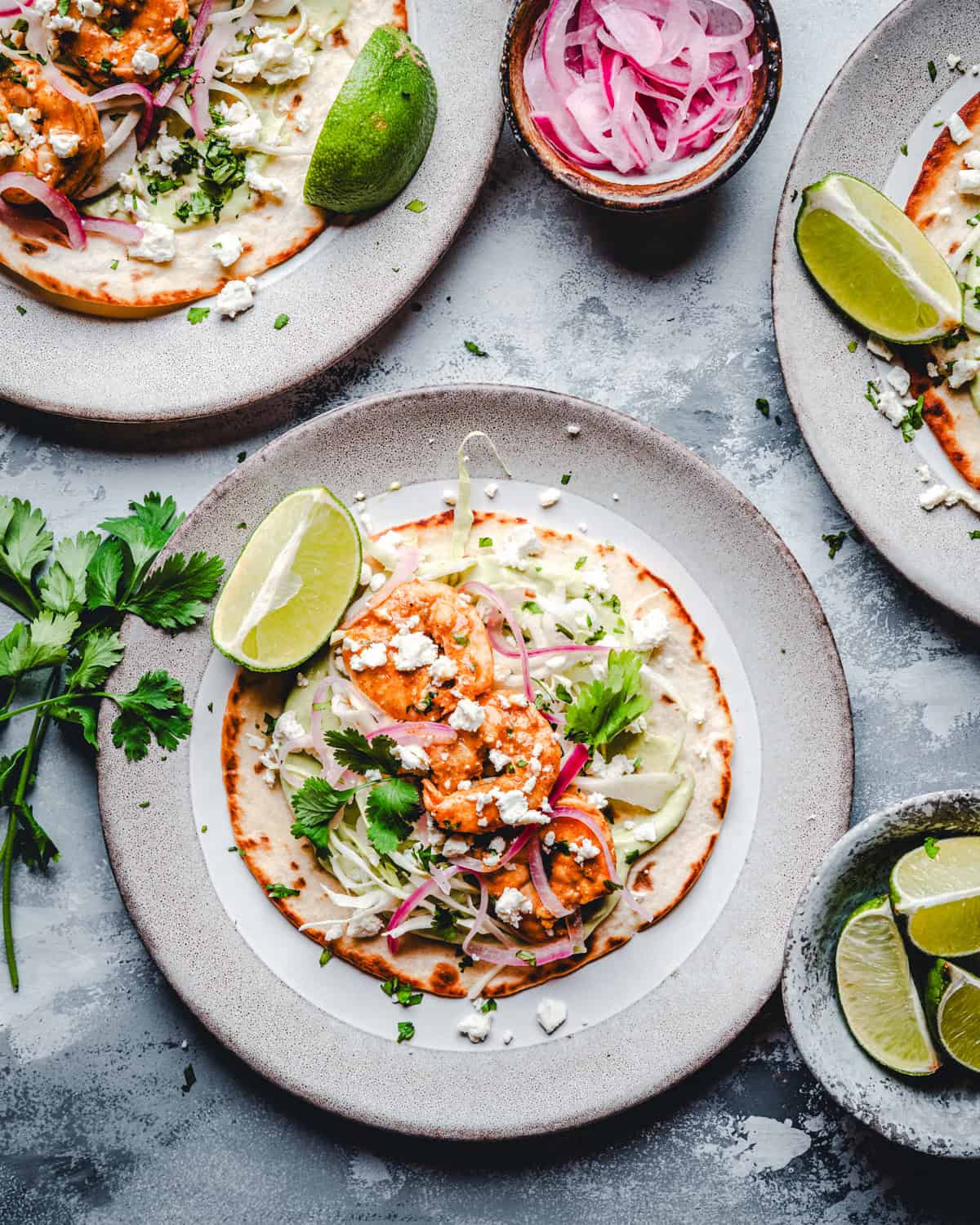 A plate with a shrimp tostada topped with shrimp, pickled onions, cilantro, crumbled cheese, and a lime wedge. Additional plates with similar tacos, lime wedges, and a bowl of pickled onions are nearby on a grey surface.