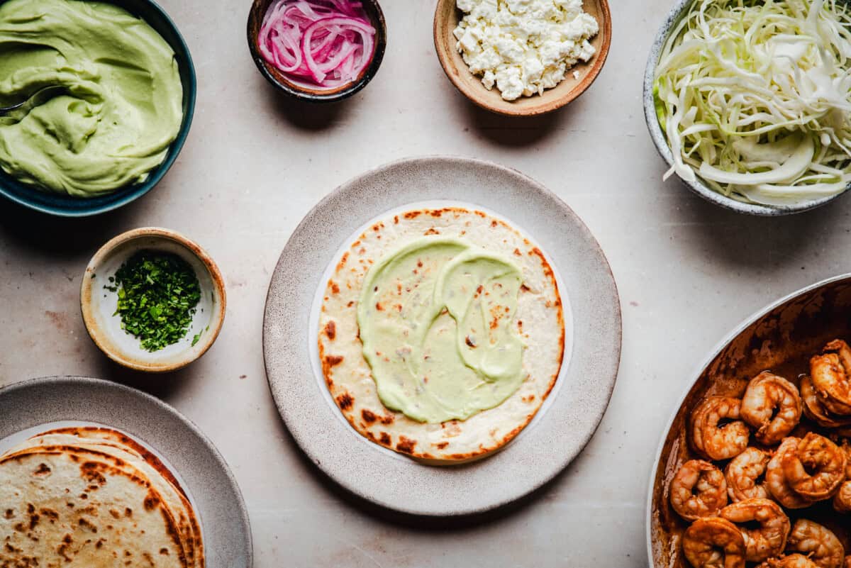 A tostada on a plate is spread with creamy avocado sauce, surrounded by bowls of pickled onions, crumbled cheese, shredded cabbage, cilantro, another tortilla, and a skillet of cooked shrimp.