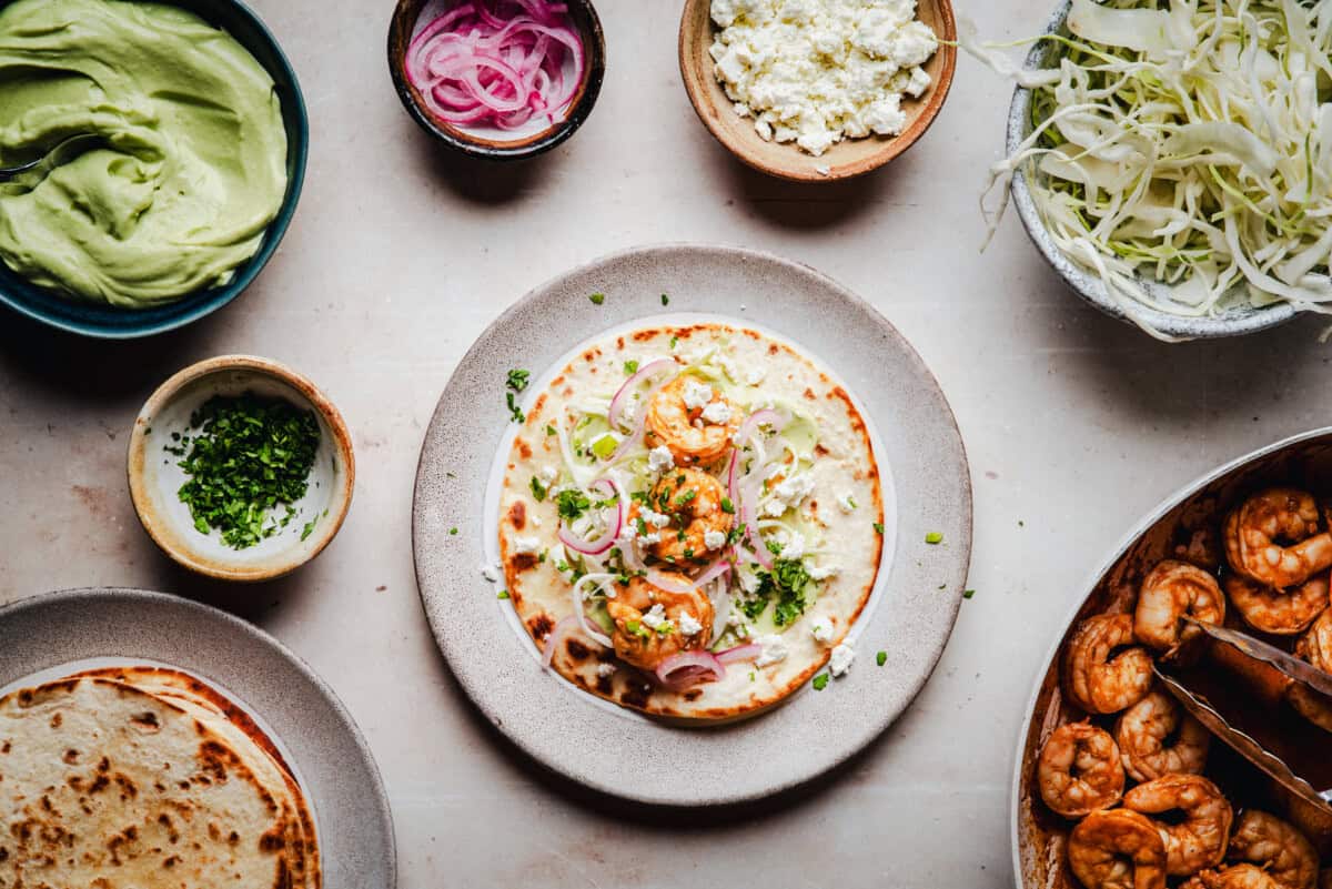 A plate with a shrimp tostada topped with pickled onions and herbs, surrounded by bowls of guacamole, pickled onions, cheese, shredded cabbage, chopped herbs, and cooked shrimp on a light surface.
