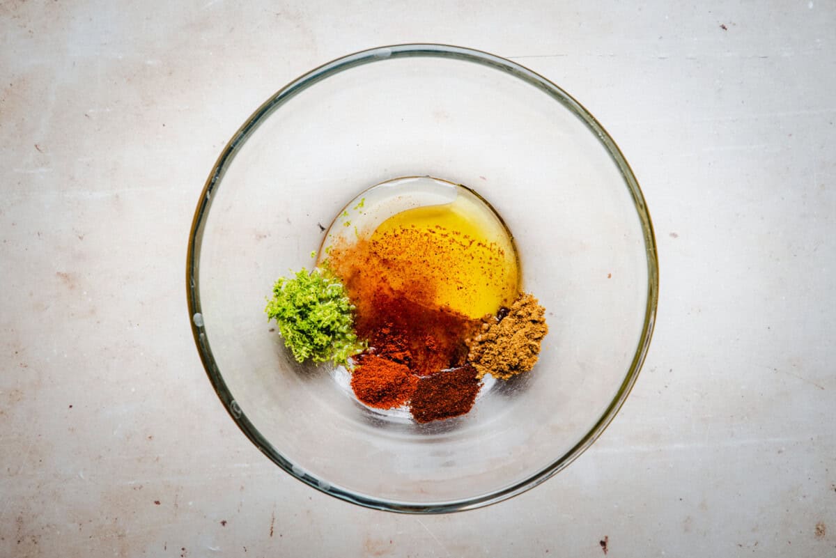 A glass bowl contains oil, ground cumin, paprika, chili powder, lime zest, and oil, arranged separately on the bottom of the bowl, displayed on a light surface.