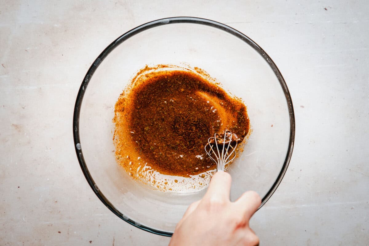 A hand uses a whisk to mix a spicy marinade in a clear glass bowl on a light-colored surface.