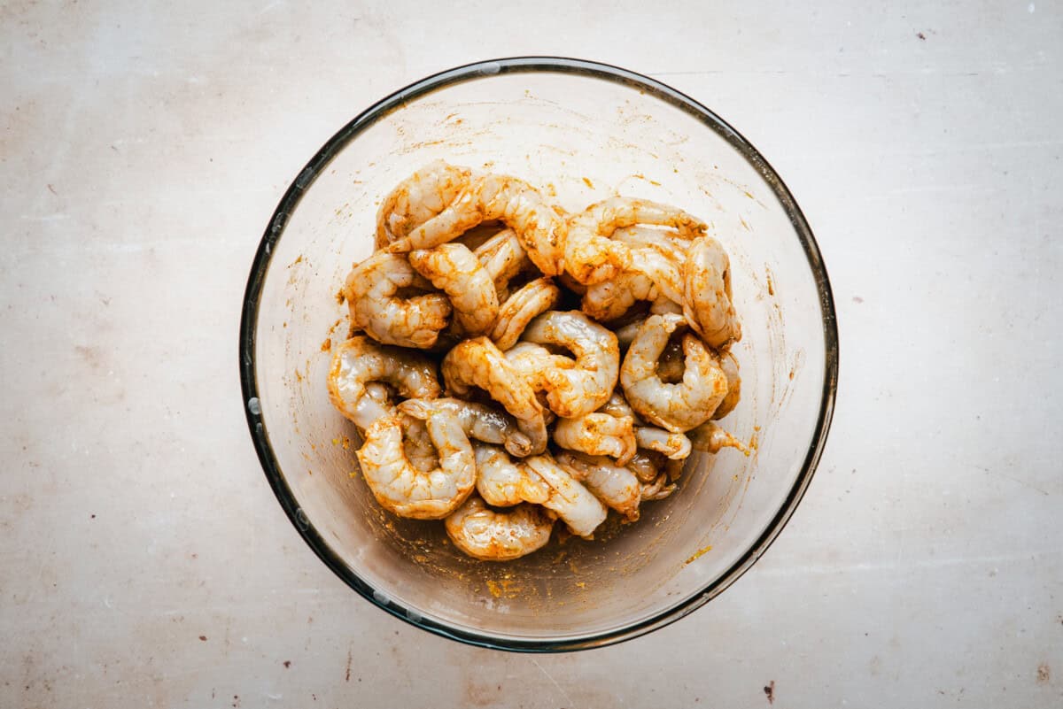 A glass bowl filled with raw shrimp marinated in spices sits on a light-colored countertop.