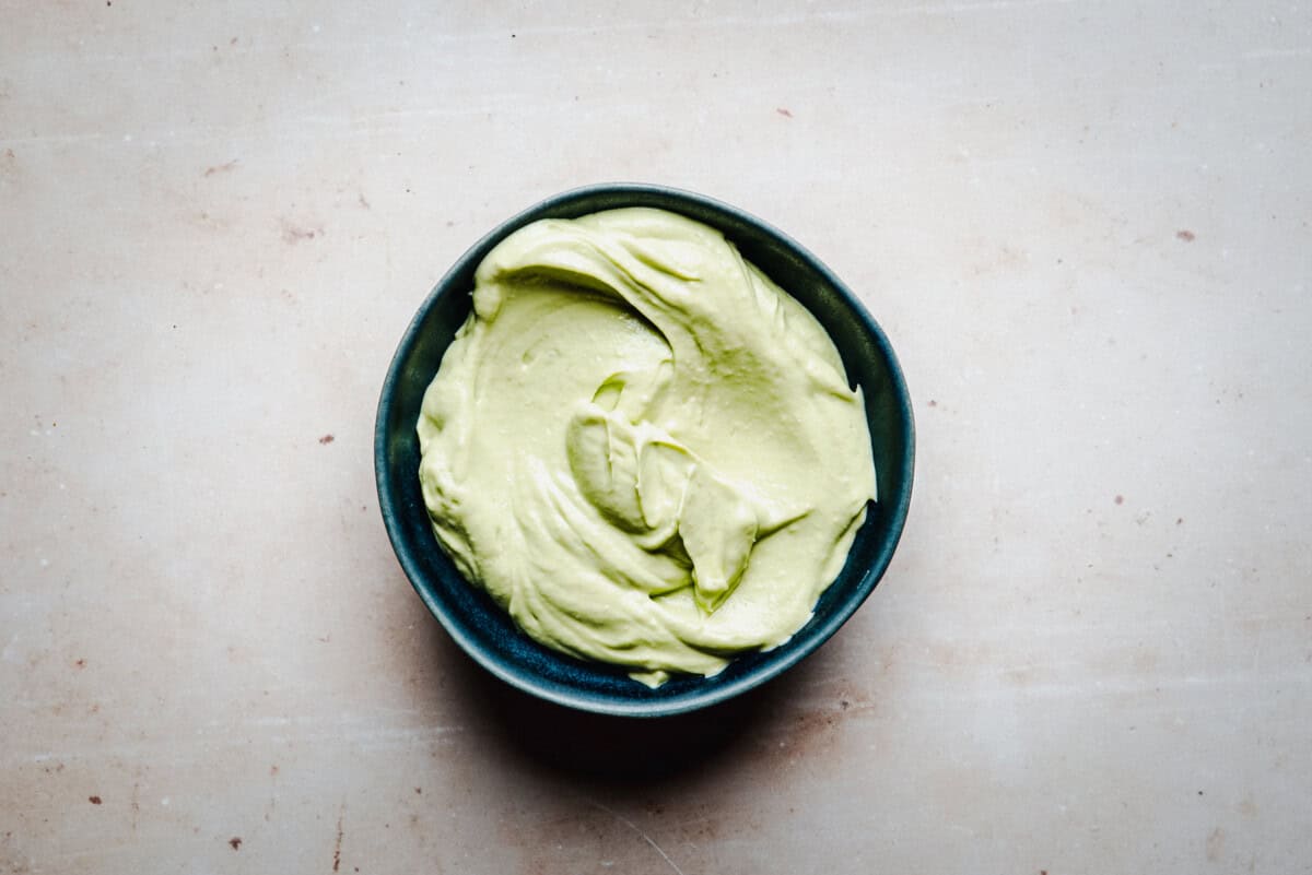 A dark bowl filled with smooth, creamy green avocado crema, placed on a light beige surface.