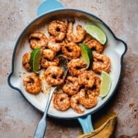 Cooked chili lime shrimp in a white skillet with a blue handle, garnished with lime wedges, on a brown textured surface. A metal spoon and a yellow cloth are also visible.