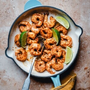 Cooked chili lime shrimp in a white skillet with a blue handle, garnished with lime wedges, on a brown textured surface. A metal spoon and a yellow cloth are also visible.
