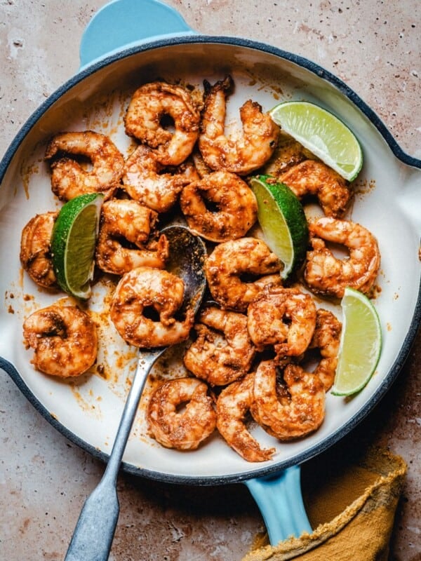 Cooked chili lime shrimp in a white skillet with a blue handle, garnished with lime wedges, on a brown textured surface. A metal spoon and a yellow cloth are also visible.