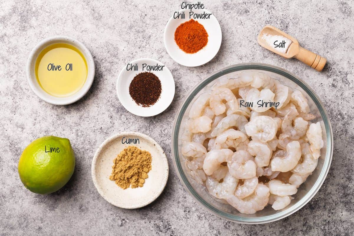 A bowl of raw shrimp surrounded by small bowls of olive oil, chili powder, chipotle chili powder, cumin, salt, and a whole lime on a gray surface. Each ingredient is labeled with handwritten text.