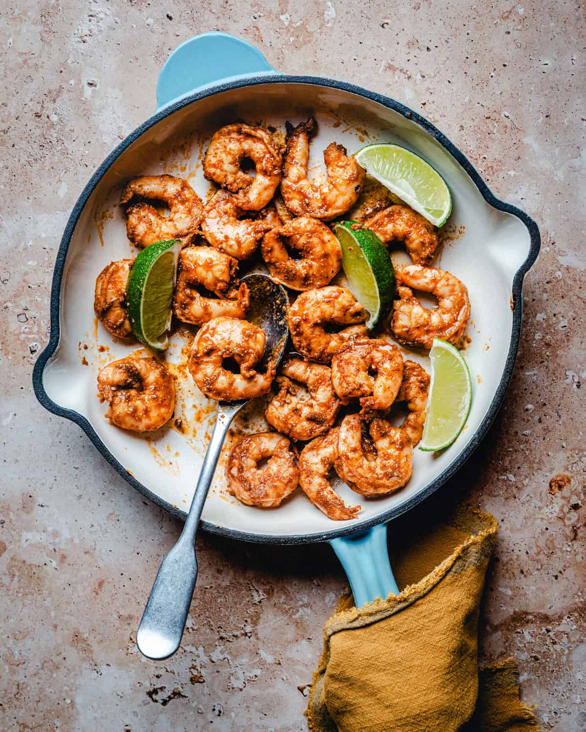 A skillet filled with cooked, chili lime shrimp garnished with lime wedges. A metal fork rests in the pan, and a yellow cloth is underneath the skillet handle. The background is a textured tan surface.