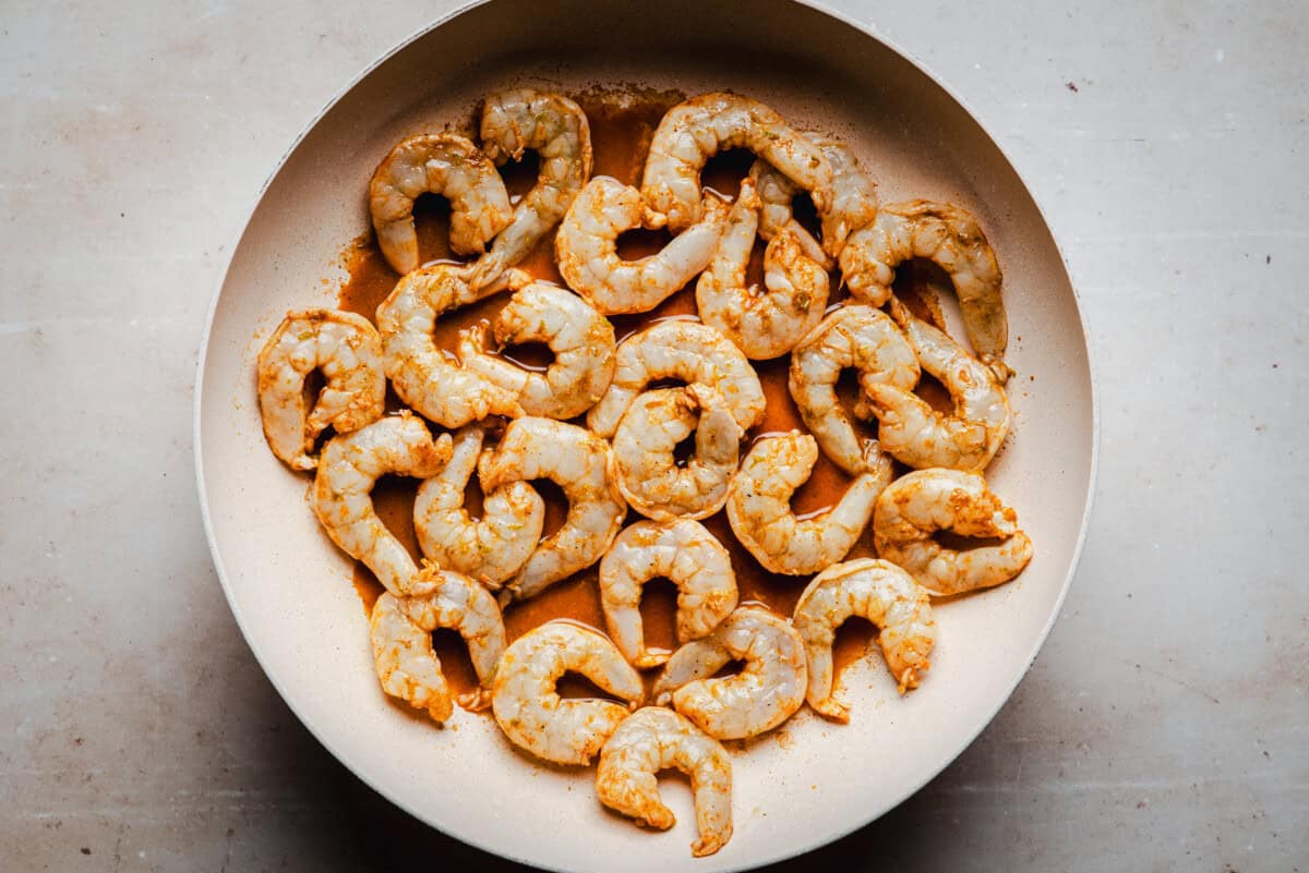 Raw shrimp marinated in spices are arranged in a single layer in a light-colored frying pan on a beige surface, ready to be cooked.