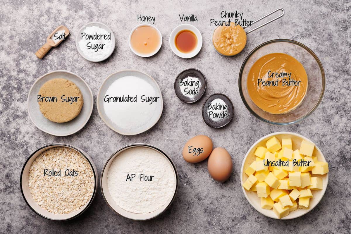 Ingredients for baking cookies, including salt, powdered sugar, honey, vanilla, chunky and creamy peanut butter, brown and granulated sugar, baking soda, baking powder, rolled oats, flour, eggs, and cubed unsalted butter.