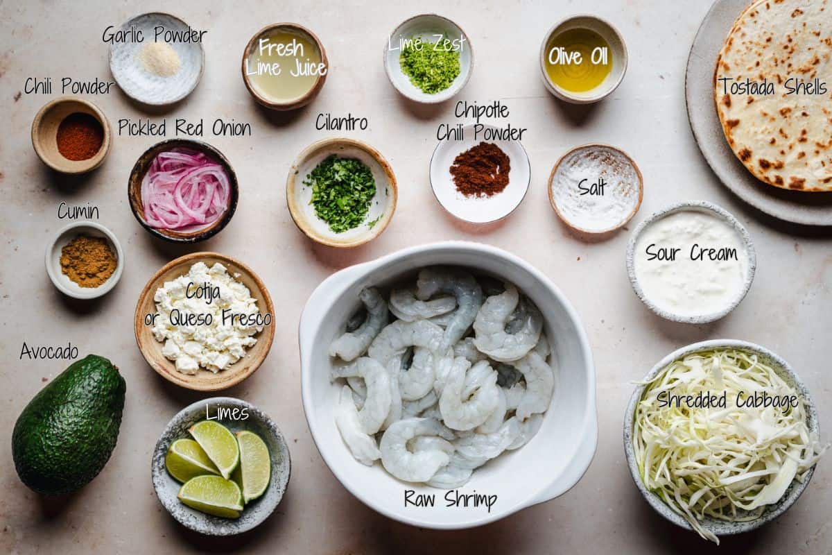 Overhead shot of shrimp tostada ingredients in bowls: raw shrimp, spices, lime, cilantro, oil, avocado, pickled onions, cheese, sour cream, salt, shredded cabbage, and tostada shells on a light background.